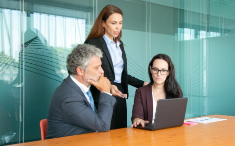 Três pessoas em uma mesa em um espaço coworking em BH. Seno um homem sentado vestindo um terno cinza, uma mulher em pé com terno preto e uma sentada em frente ao notebook de terno marrom e óculos.