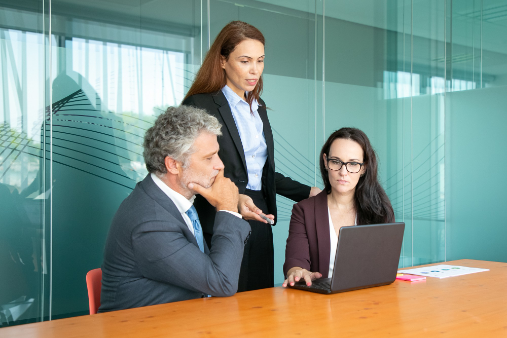 Três pessoas em uma mesa em um espaço coworking em BH. Seno um homem sentado vestindo um terno cinza, uma mulher em pé com terno preto e uma sentada em frente ao notebook de terno marrom e óculos.